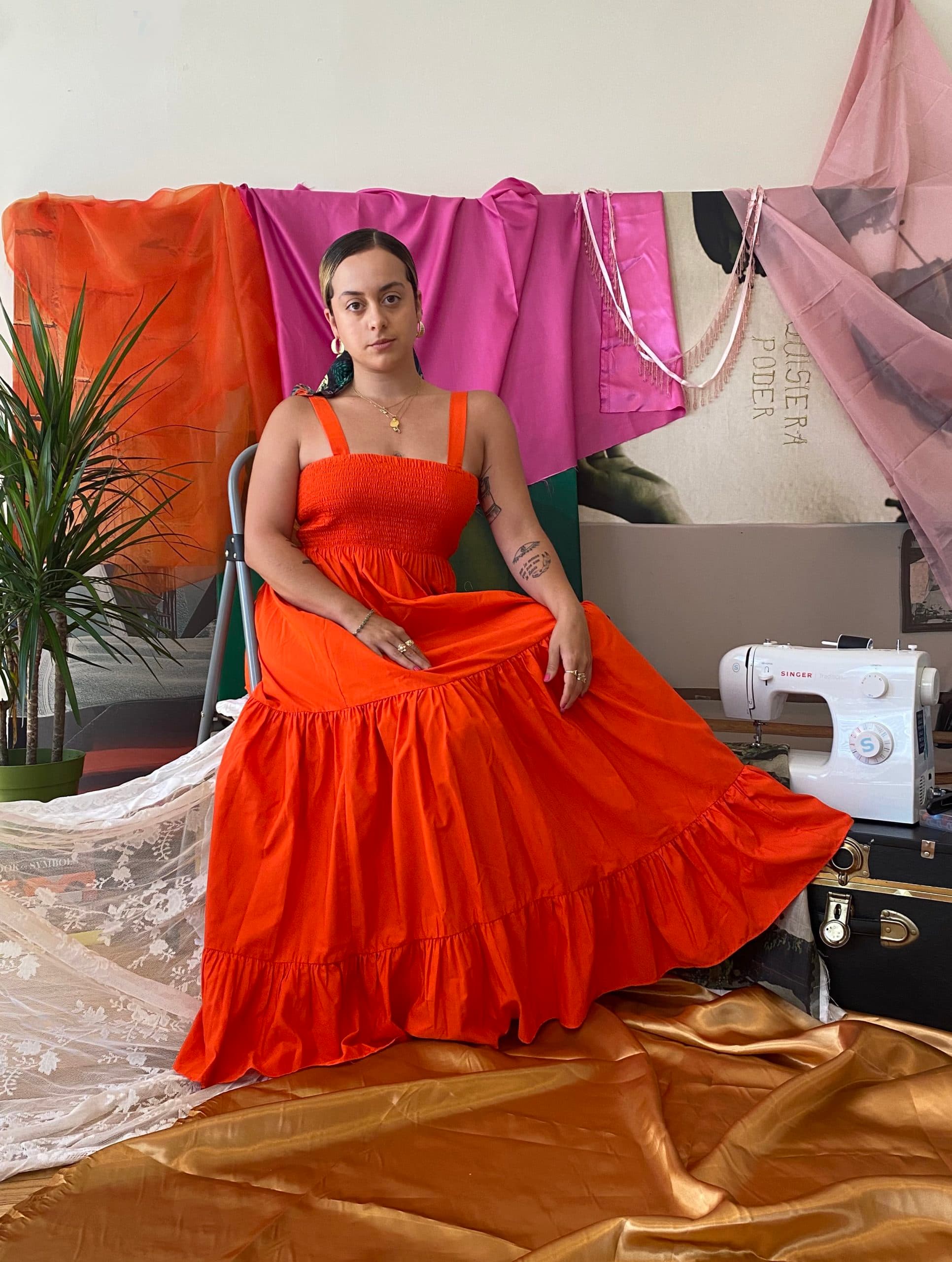 A portrait of Paola de la Calle wearing a bright red dress, seated in front of displays of fabric.