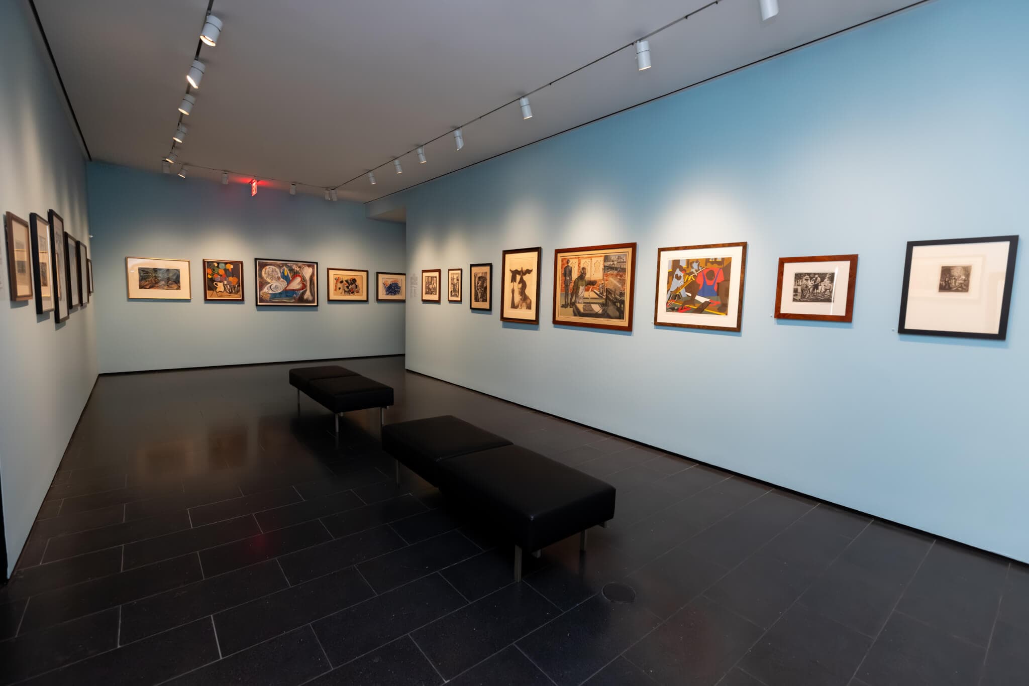 Framed artworks hang on the powder blue wall of a gallery.