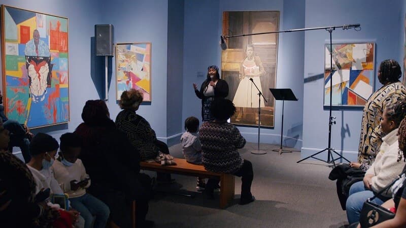 Singer Ashley Villard performs in front of an audience in a room that displays artwork on the walls.
